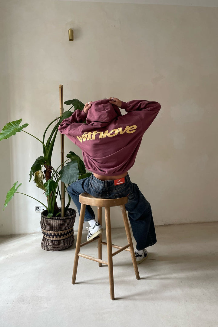 Bordeauxroter Oversized-Hoodie mit gelbem „withlove“-Rückendruck, Kapuze und lockerer Passform auf einem Barhocker.
