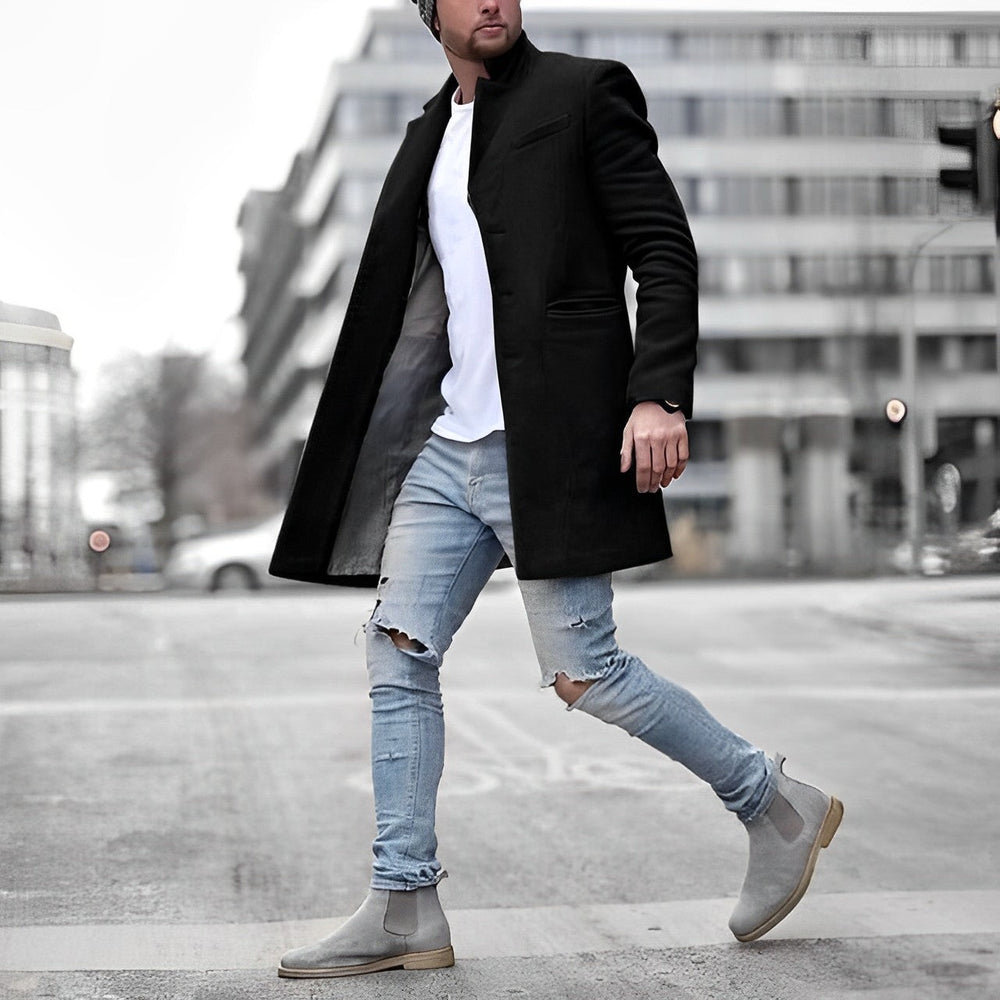 Schwarzer Slim-Fit Herrenmantel mit Stehkragen, kombiniert mit weißem Shirt, hellblauen Ripped-Jeans und grauen Chelsea Boots.