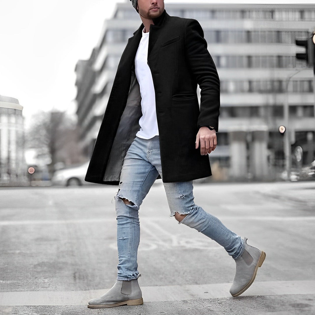 Schwarzer Slim-Fit Herrenmantel mit Stehkragen, kombiniert mit weißem Shirt, hellblauen Ripped-Jeans und grauen Chelsea Boots.
