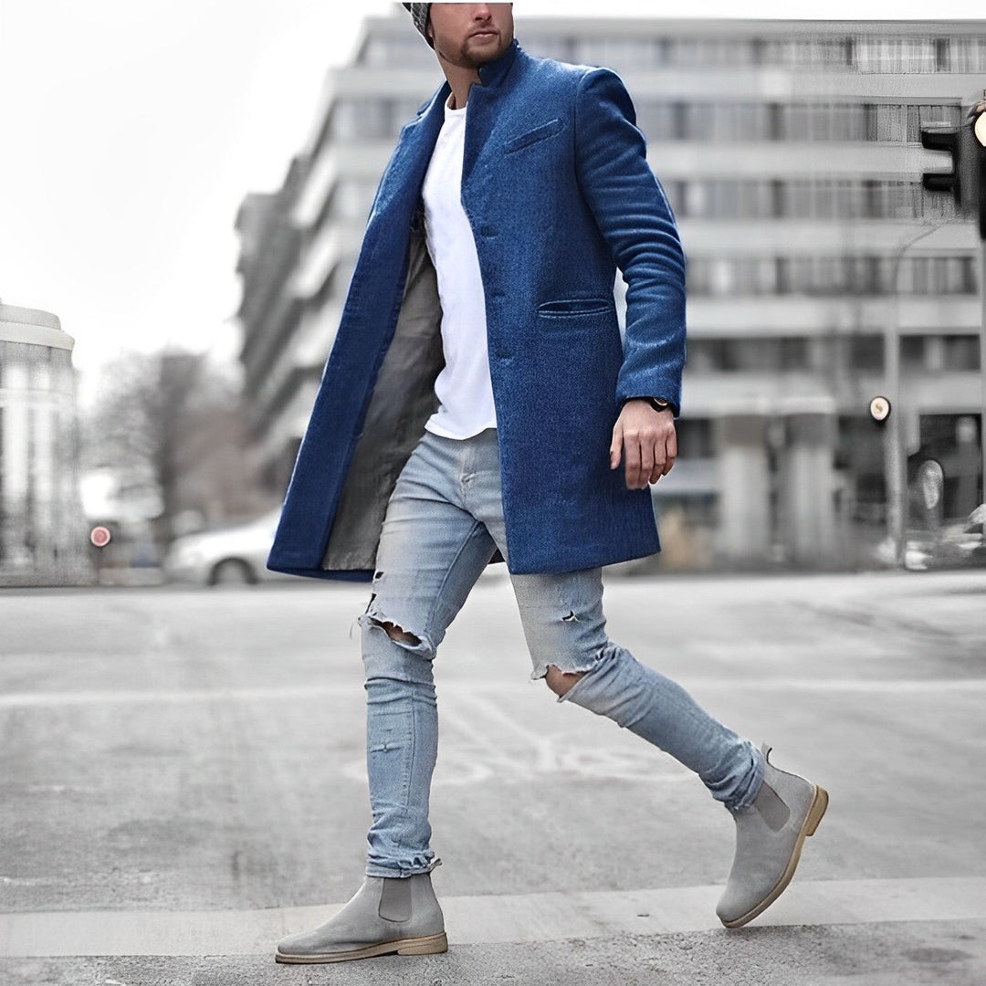 Blauer Slim-Fit Herrenmantel mit Stehkragen, kombiniert mit weißem T-Shirt, zerrissenen Jeans und grauen Chelsea Boots.