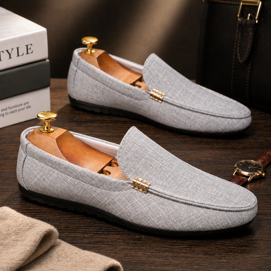 Grauer Herren-Loafer aus Canvas mit Slip-On-Design, goldfarbenem Detail und flexibler schwarzer Sohle auf dunklem Holz.