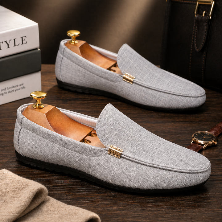 Grauer Herren-Loafer aus Canvas mit Slip-On-Design, goldfarbenem Detail und flexibler schwarzer Sohle auf dunklem Holz.
