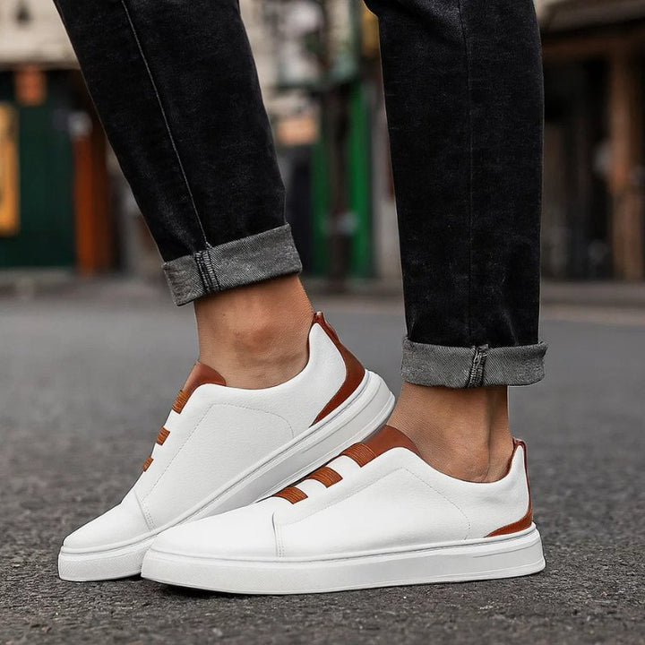 Weißer Herren-Sneaker aus Leder zum Hineinschlüpfen mit braunen elastischen Bändern und Fersendetails auf Asphalt.