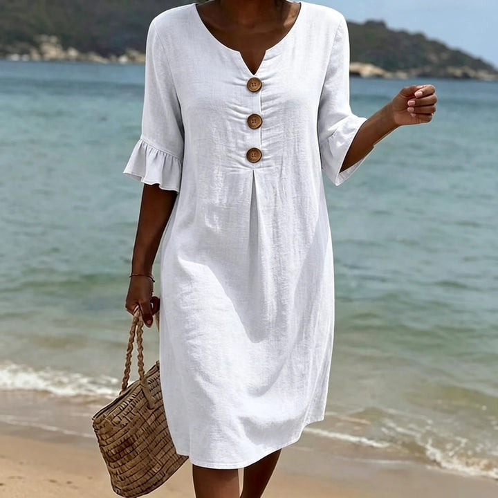 Weißes Fiona Midikleid aus Leinen mit V-Ausschnitt, Volant-Ärmeln und Holzknöpfen am Strand.