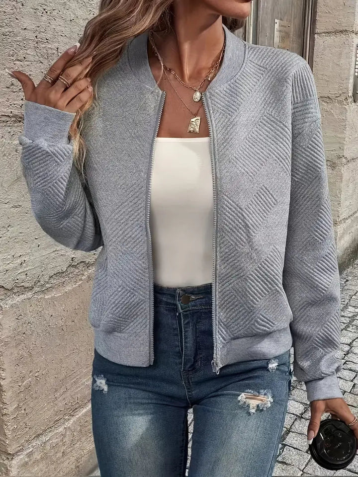 Graue Strickjacke mit Reißverschluss, Stehkragen und strukturiertem Muster, kombiniert mit Jeans und weißem Top.