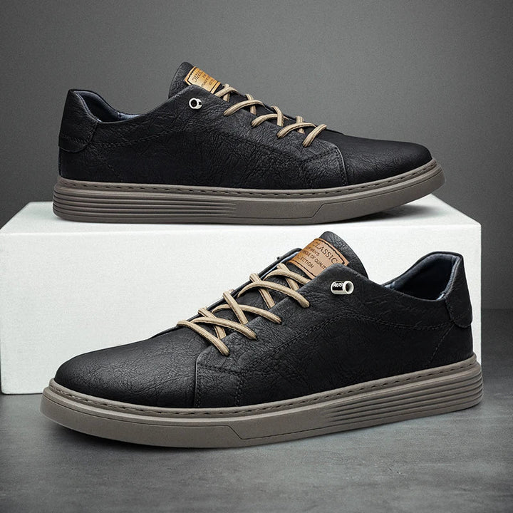 Schwarze Herren-Sneaker aus strukturiertem Leder mit beigen Schnürsenkeln und gerippter, flacher Sohle auf einem weißen Block.