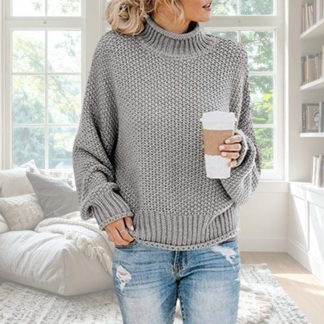 Grauer Damen-Grobstrickpullover mit Rollkragen, gerippten Bündchen und lockerer Passform in einem gemütlichen Wohnzimmer.