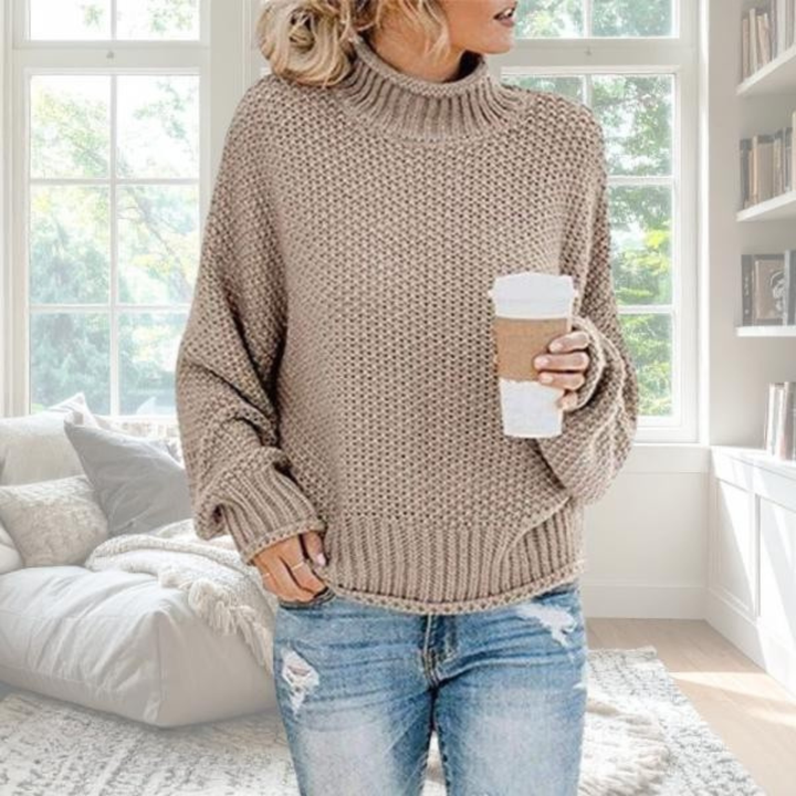Beiger Damen-Grobstrickpullover mit Rollkragen, gerippten Bündchen und lockerer Passform in einem hellen Wohnzimmer.