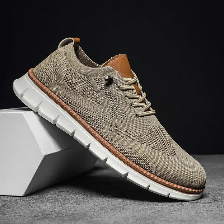 Beiger Herren-Mesh-Sneaker mit Schnürung, braunen Lederdetails und flexibler weißer Laufsohle auf einem weißen Block.