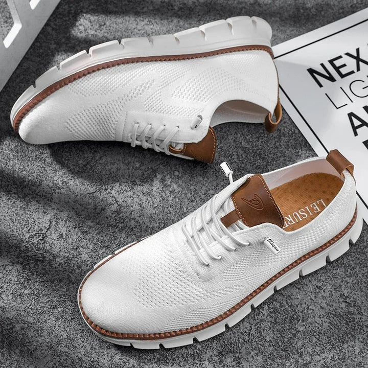 Weißer Herren-Mesh-Sneaker mit braunen Lederdetails, Schnürung und flexibler weißer Laufsohle auf grauem Untergrund.