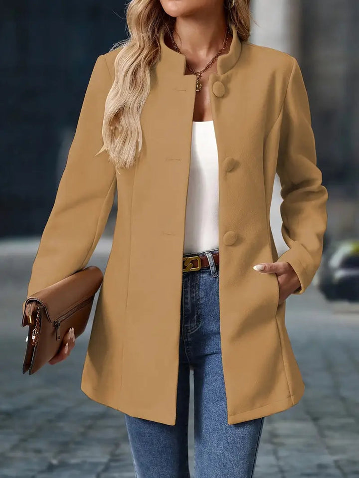 Beiger Damenmantel mit Stehkragen, Knopfleiste und tailliertem Schnitt, kombiniert mit Jeans und einer Clutch.