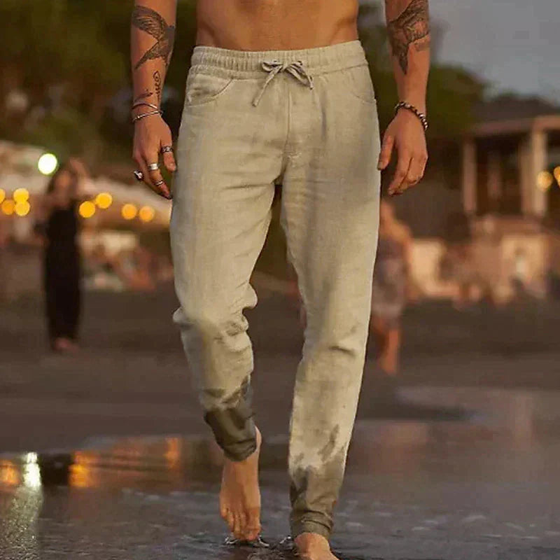 Beige Herren-Leinenhose mit elastischem Kordelzugbund im lockeren Schnitt, getragen am Strand.