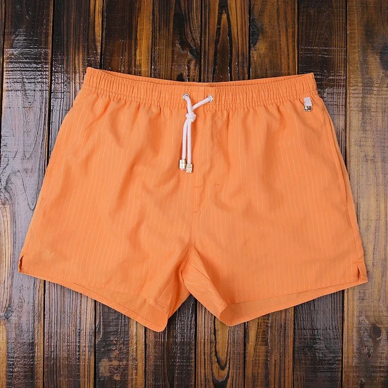 Orangefarbene Herren-Badeshorts mit feinen weißen Streifen, elastischem Bund und weißem Kordelzug auf Holzuntergrund.