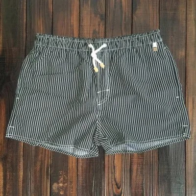 Schwarze Herren-Badeshorts mit weißen Nadelstreifen, elastischem Bund und weißem Kordelzug auf dunklem Holzuntergrund.
