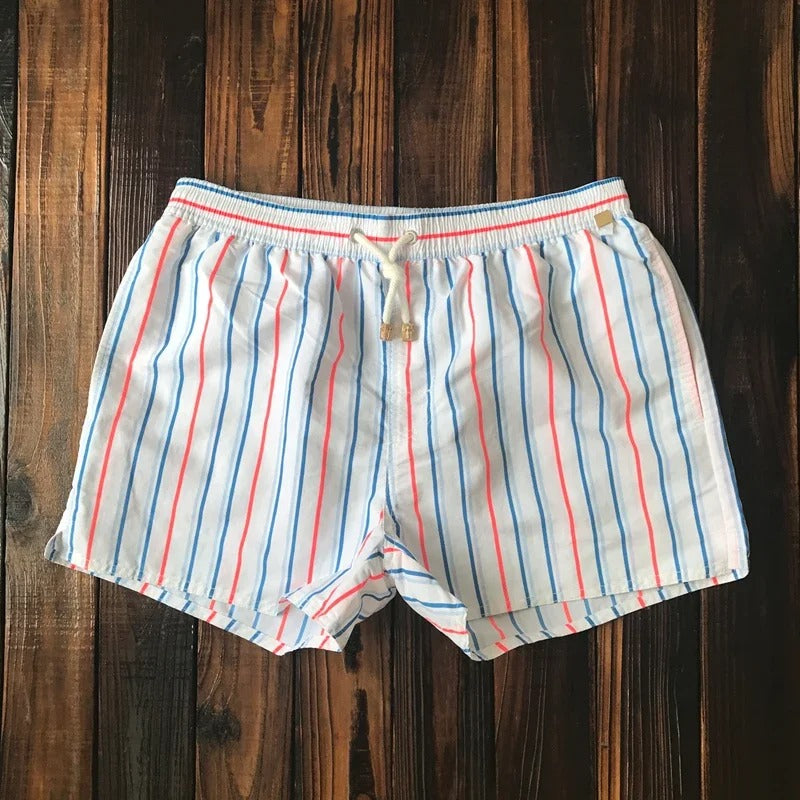 Lukas Herren-Badeshorts mit blau-rotem Streifenmuster, elastischem Bund und weißem Kordelzug auf dunklem Holzuntergrund.