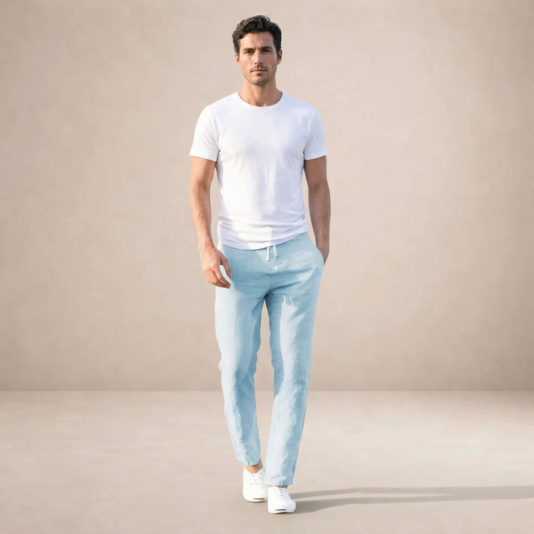 Mann trägt eine hellblaue, knöchellange Leinenhose mit Kordelzug und weißem T-Shirt vor beigem Hintergrund.