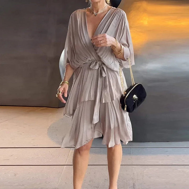 Beiges Midikleid mit V-Ausschnitt, Wickeloptik, Taillenband und gestuftem Rock aus fließendem Stoff.