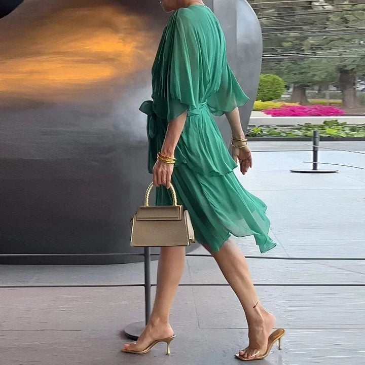 Grünes Midikleid mit V-Ausschnitt, Wickeloptik, Taillenbindung und gestuftem Rock, kombiniert mit goldener Handtasche.