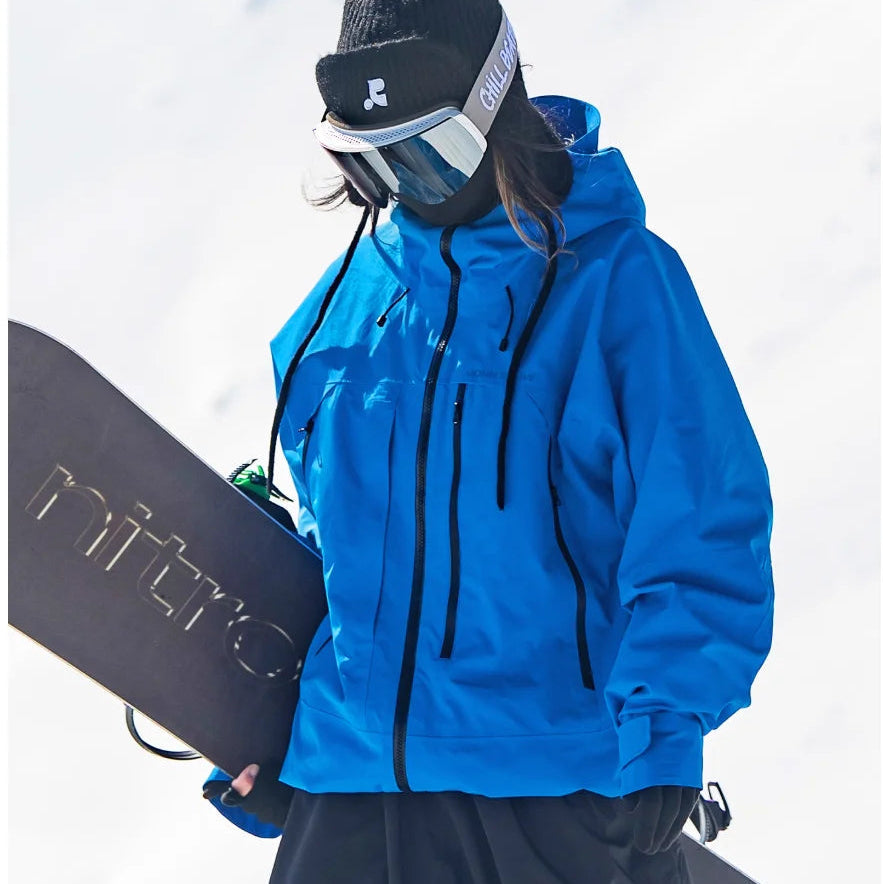 Blaue, wasserdichte Melina Damen-Skijacke mit Kapuze und Reißverschlusstaschen beim Snowboarden im Schnee.