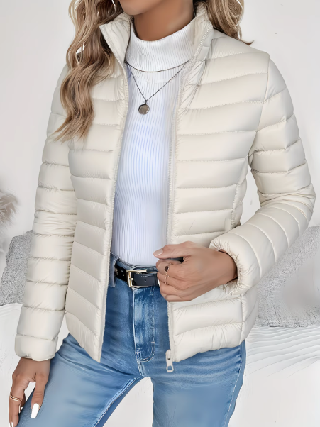 Taillierte, beige Miriam Steppjacke mit Stehkragen und Reißverschluss, kombiniert mit weißem Rollkragenpullover und Jeans.
