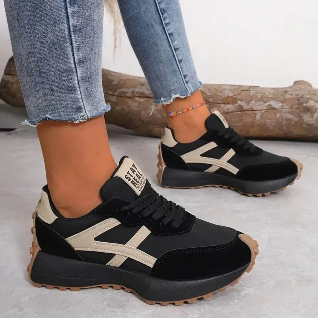 Schwarze Retro-Sneaker mit beigen Akzenten, Schnürung, markantem Seitenstreifen und profilierter Gummisohle an Damenfüßen.
