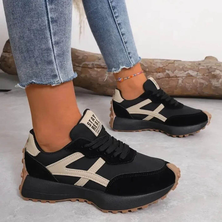 Schwarze Retro-Sneaker mit beigen Akzenten, Schnürung, markantem Seitenstreifen und profilierter Gummisohle an Damenfüßen.