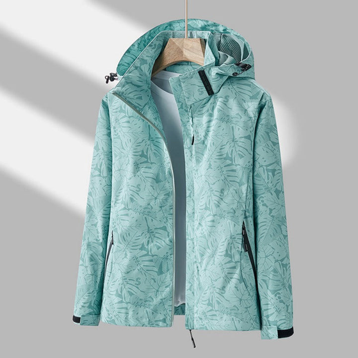 Wasserdichte Damenjacke in Mintgrün mit Blättermuster, Kapuze und Reißverschlusstaschen an einem Kleiderbügel.