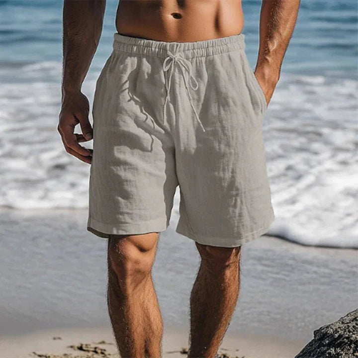 Beige Herren-Leinenshorts mit Kordelzug, elastischem Bund und Seitentaschen in lockerer Passform am Strand.