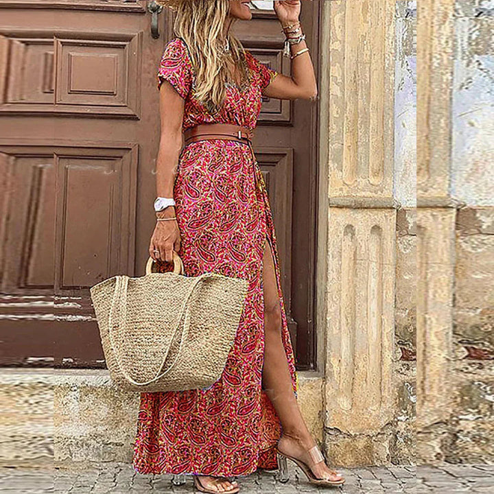Buntes Boho-Maxikleid mit V-Ausschnitt, kurzem Ärmel, Beinschlitz und braunem Gürtel, kombiniert mit einer Strohtasche.