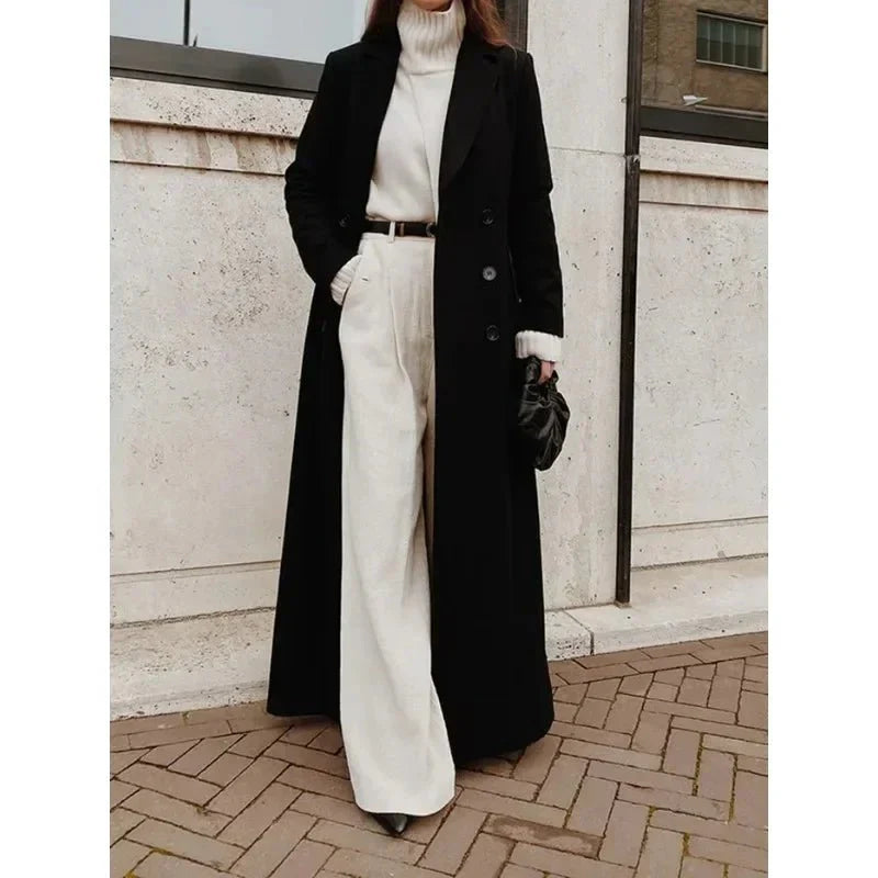 Schwarzer, bodenlanger Damenmantel mit zweireihiger Knopfleiste, breitem Revers und Taschen über weißem Outfit.