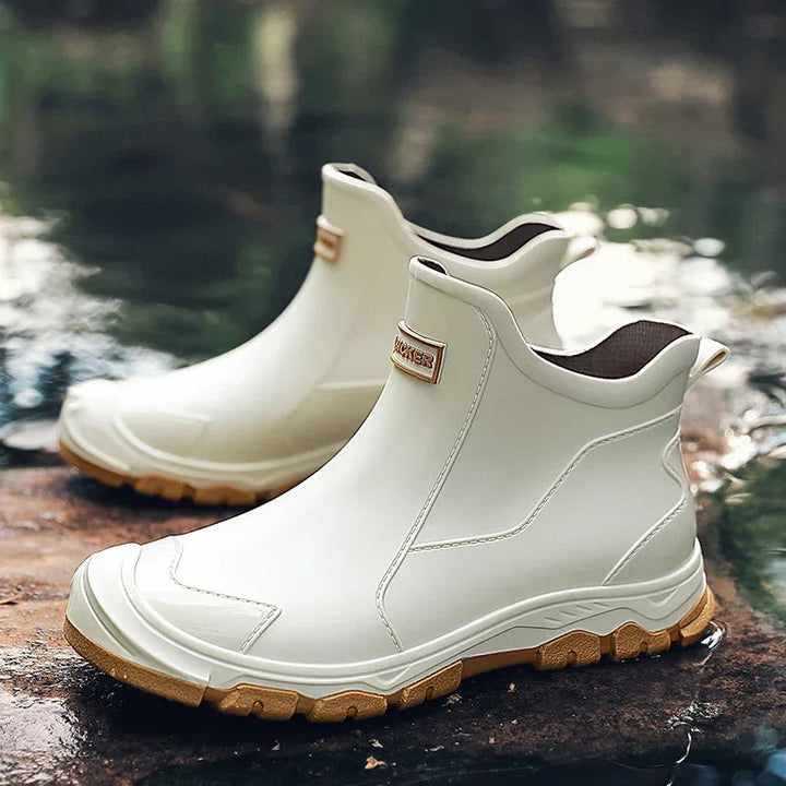 Weiße, wasserdichte Damen-Regenstiefeletten mit Slip-on-Design, verstärkter Zehenkappe und brauner Profilsohle auf nassem Boden.