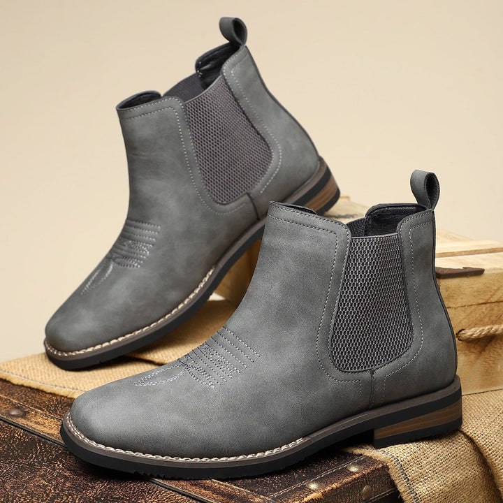 Graue Herren-Chelsea-Boots aus Kunstleder mit Western-Nähten, elastischen Einsätzen und Blockabsatz auf einer Holzkiste.