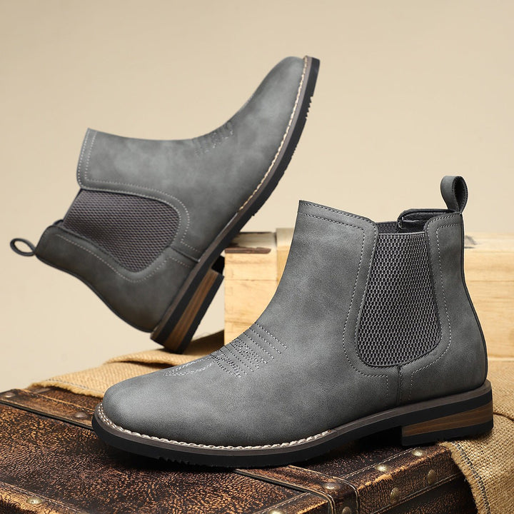 Graue Herren Chelsea Boots in Lederoptik mit Western-Nähten, elastischen Einsätzen und Blockabsatz auf einer Holzkiste.