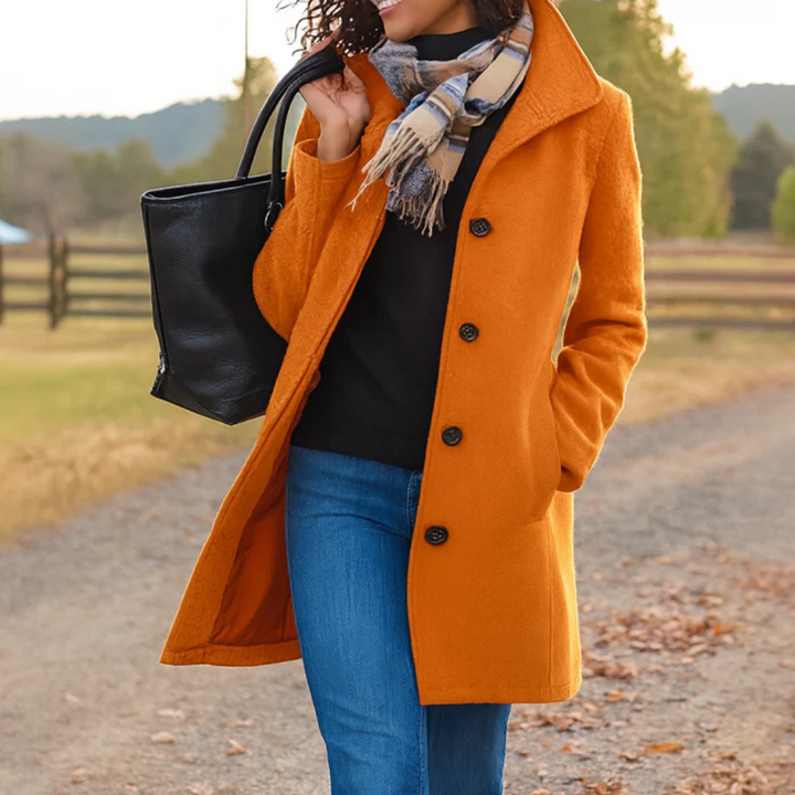 Eleganter, orangefarbener Damen-Wintermantel mit Knopfleiste, Umlegekragen und Seitentaschen in herbstlicher Umgebung.